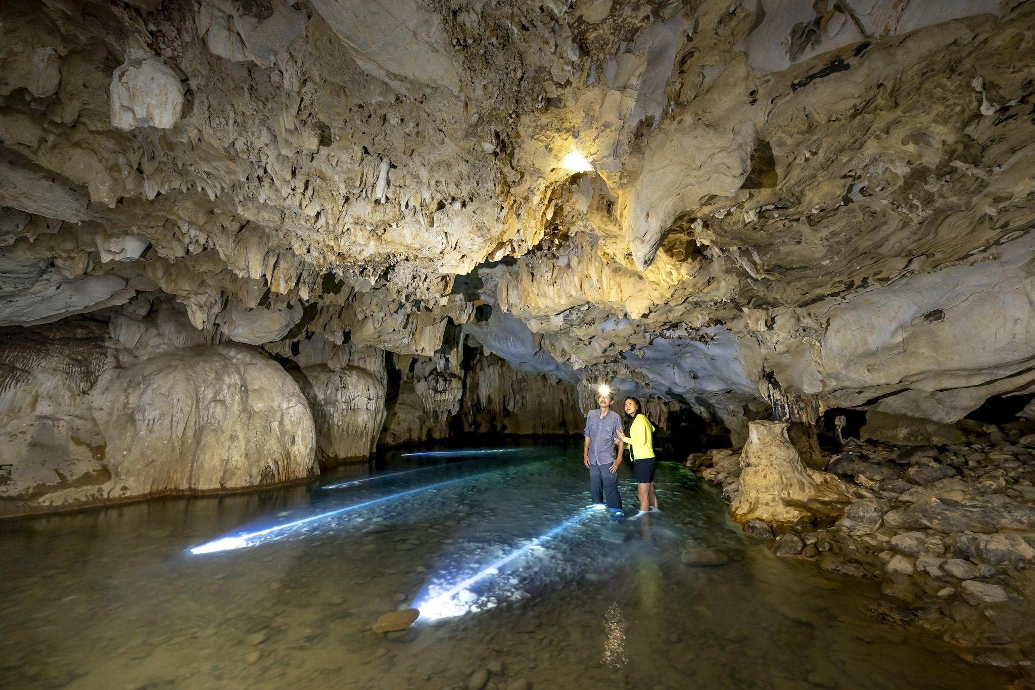 Two people exploring a beautifully illuminated underground cave with water reflections.